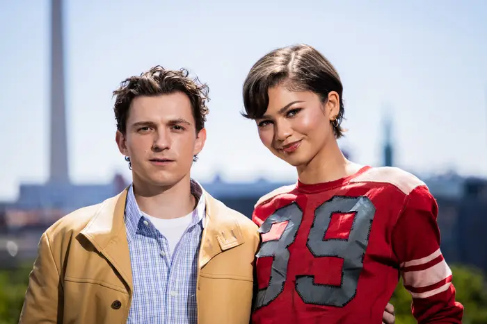 Zendaya og Tom står utendørs. Personen til venstre har på seg en uformell jakke og skjorte. Personen til høyre har på seg en rød topp med "39" på den
