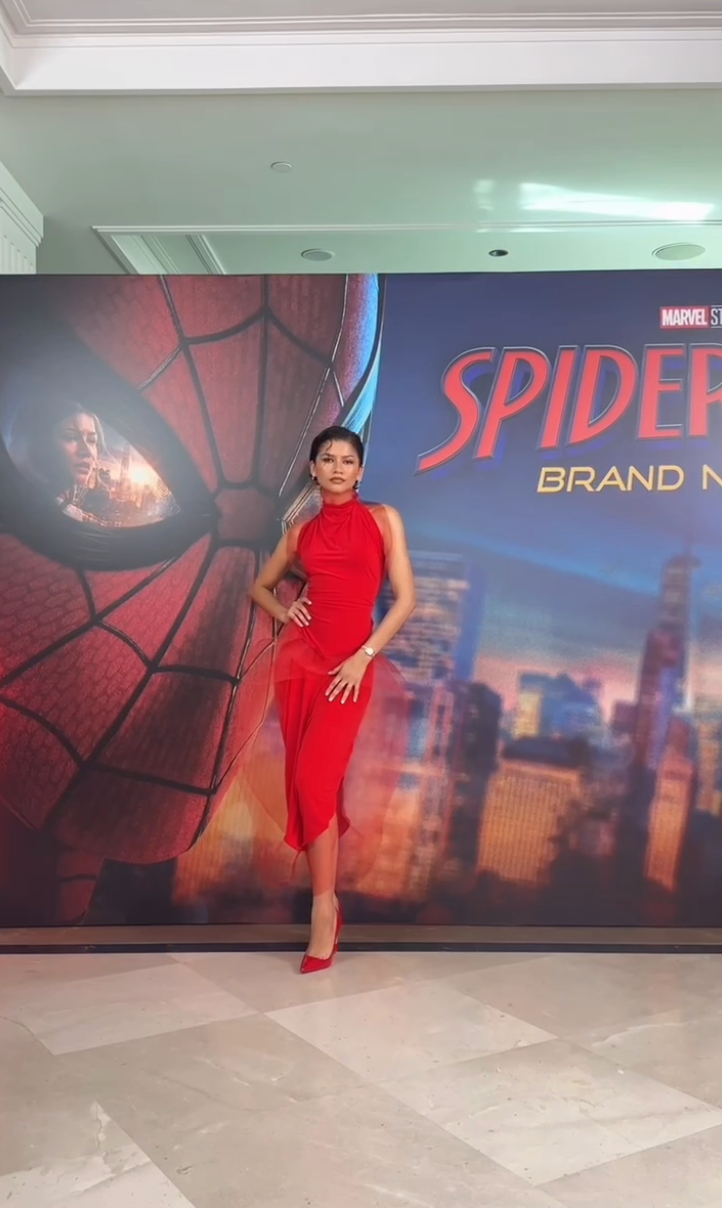 Zendaya poserer i en elegant, figursydd rød kjole og hæler foran en "Spider-Man: Brand New" plakat på et arrangement