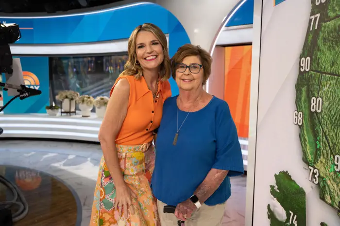 To kvinner som smiler i et TV-studio; den ene i oransje topp med blomsterskjørt, den andre i blå bluse. De poserer ved siden av en stor kartvisning