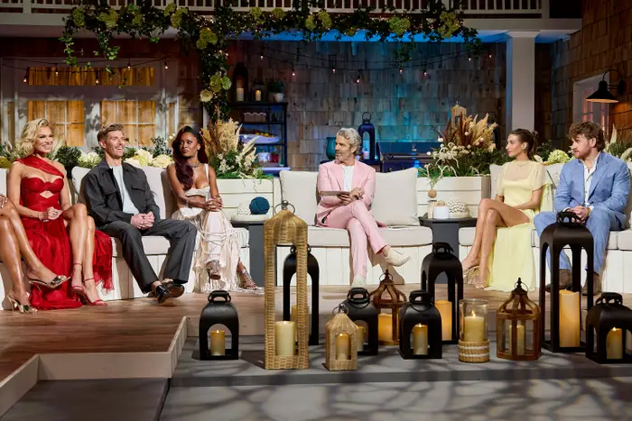 En gjenforeningsscene med TV-show med seks personer sittende. Deltakerne bruker formell antrekk, inkludert kjoler og dresser. Stearinlys og lykter pryder settet