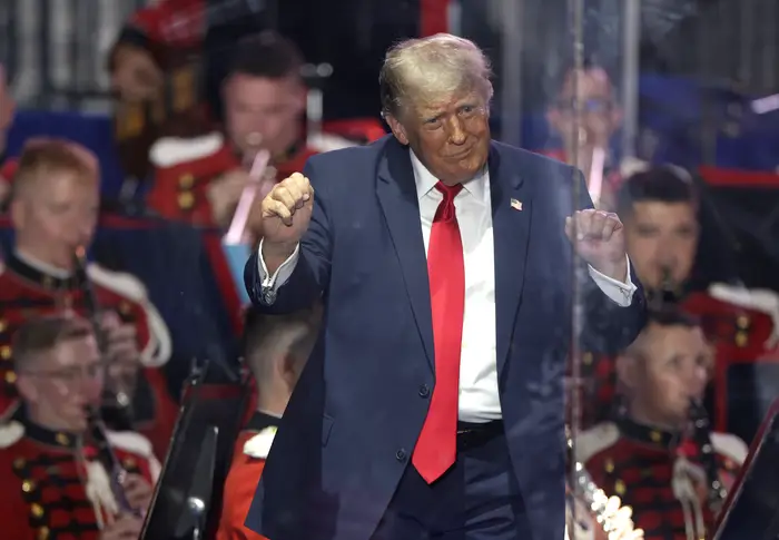 Donald Trump gestikulerer energisk foran et band i uniform. De står bak en tydelig barriere på en scene