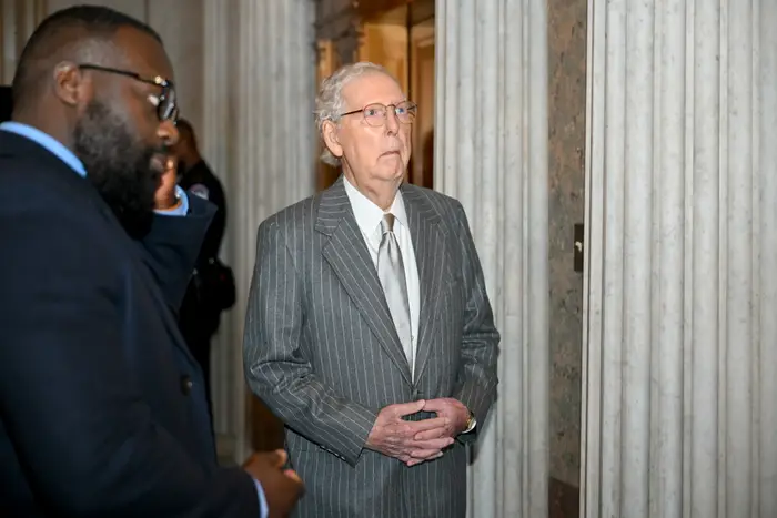 En eldre mann i nålestripet drakt står innendørs, omgitt av andre