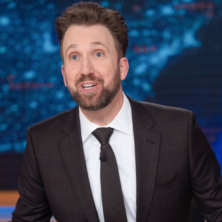 En person i dress sitter ved "The Daily Show"-pulten på settet, gestikulerer med begge hender, foran et grafisk bakteppe