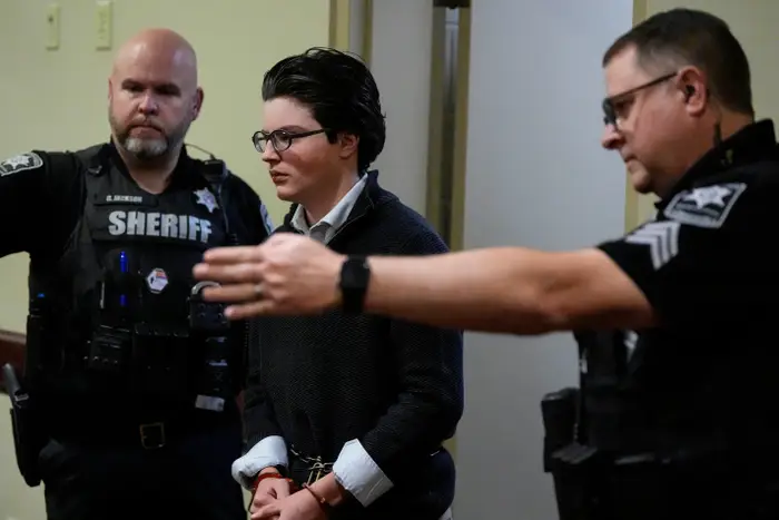 En person i håndjern, eskortert av to offiserer, står i en gang. De har på seg en genser over en skjorte med krage og briller