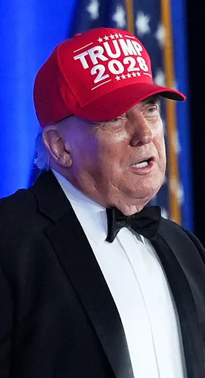 En mann i smoking og "Trump 2024" hatten snakker på et podium. En sittende kvinne lytter; et amerikansk presidentstempel er på podiet