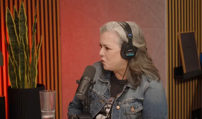 Person med hodetelefoner som snakker inn i en mikrofon, sittende i et podcaststudio. En plante er synlig i bakgrunnen