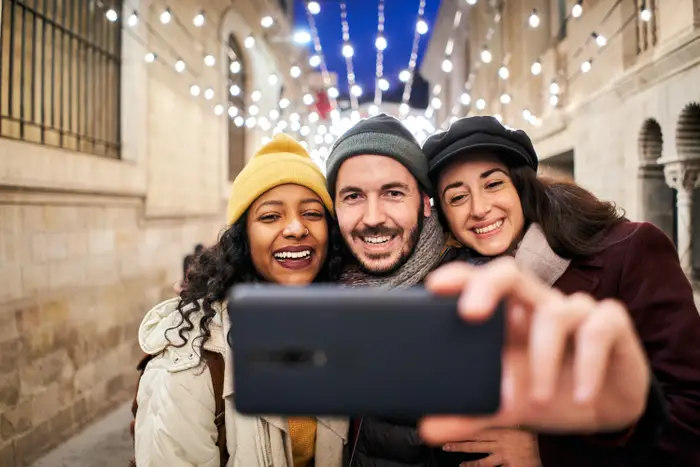 Tre venner i vinterklær tar en selfie under lysslynge i en bygate, smiler og nyter øyeblikket