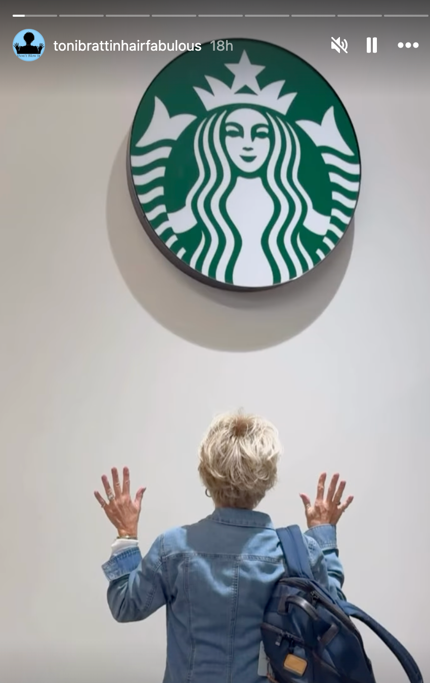 Person med kort hår og dongerijakke løfter hendene mot en stor Starbucks-logo på veggen over