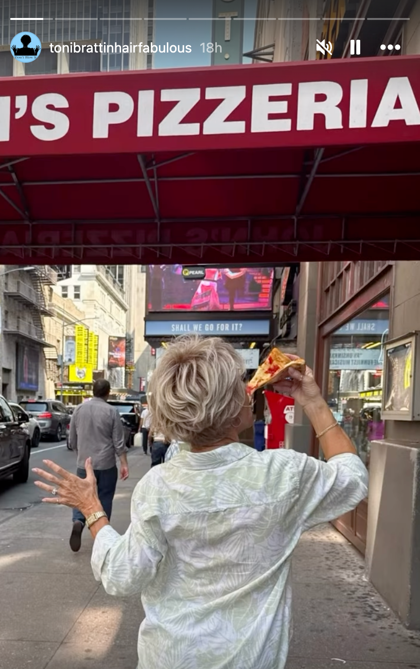 En person med kort hår går under et pizzeriaskilt, holder en pizzaskive og rekker opp den andre hånden, med en travel bygate i bakgrunnen
