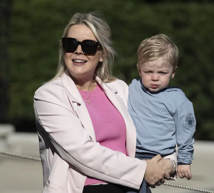 En kvinne i solbriller og en stilig blazer smiler mens hun holder et lite barn i et uformelt antrekk utendørs
