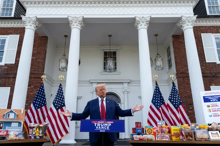 En offentlig person taler ved et podium foran en bygning med søyler, omgitt av amerikanske flagg og bord som viser ulike matvarer