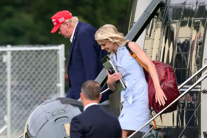 En person i dress og rød caps, og en kvinne i en ermeløs kjole som bærer en rød bag, går ut av et helikopter med en annen person i dress i nærheten