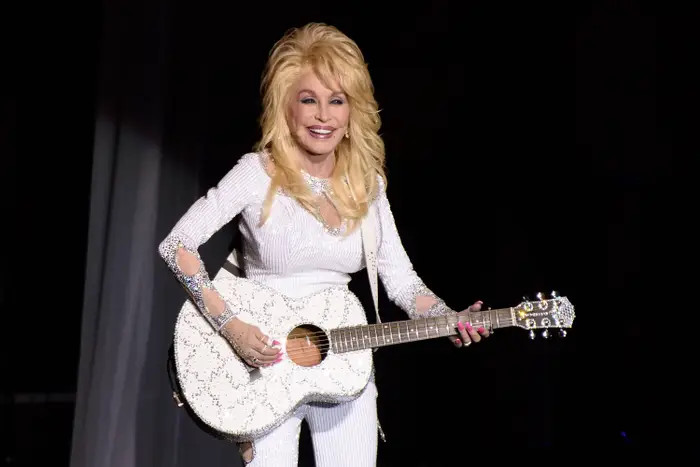 En kvinne opptrer på scenen med en hvit akustisk gitar, iført et glitrende antrekk pyntet med rhinestones