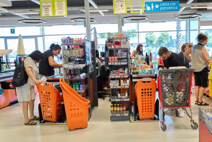 Folk handler i en matbutikk, med vogner og kurver fylt med forskjellige varer. Hyllene er fylt med mat og drikke