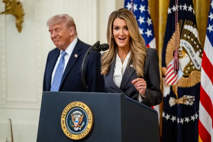 En kvinne i blazer snakker på et podium med presidentseglet, mens en mann står og smiler i bakgrunnen