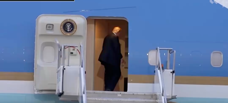 En person i dress og slips går inn i et fly via en trapp som fører til en dør