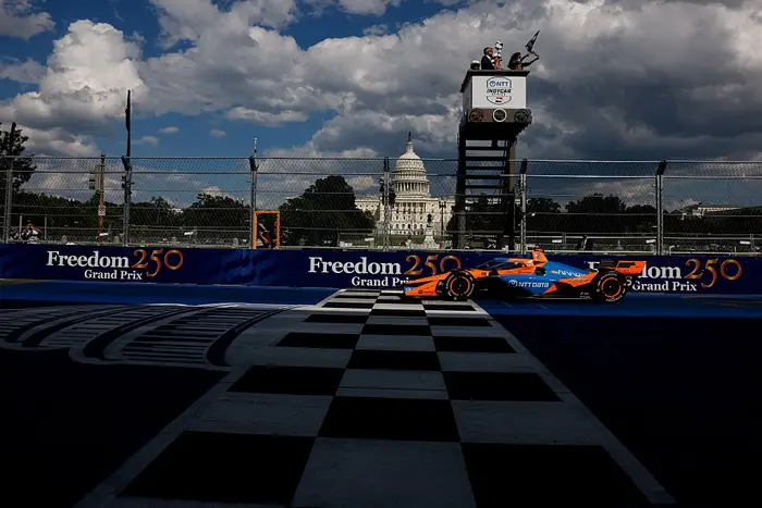 Racerbil krysser målstreken ved Freedom 250 Grand Prix, med funksjonærer i bakgrunnen og Capitol-bygningen synlig i det fjerne