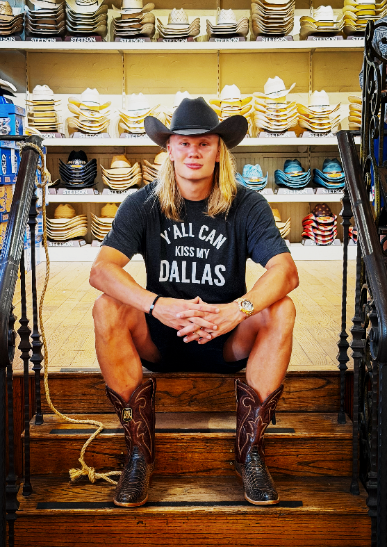 Person som har på seg cowboyhatt, "Y'ALL CAN KISS MY DALLAS" skjorte, og cowboystøvler sitter på tretrapper, omgitt av hatter i hyllene
