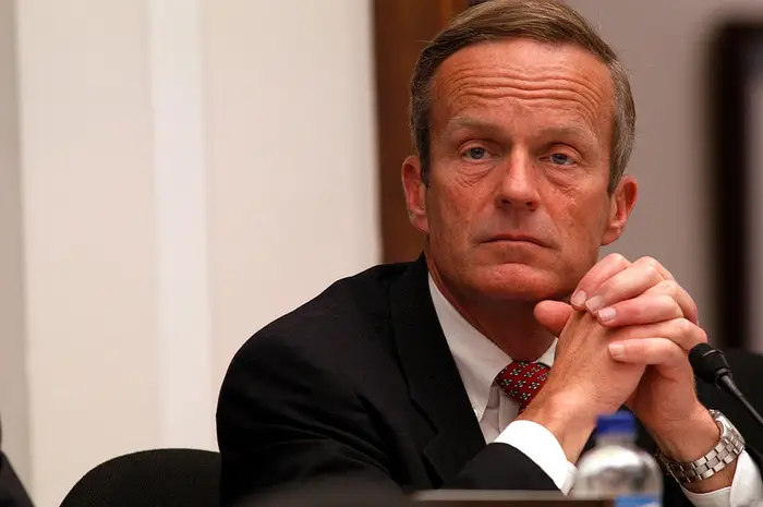 Todd Akin i dress sittende ved et bord, hendene sammen, virker gjennomtenkt under en formell begivenhet eller et møte