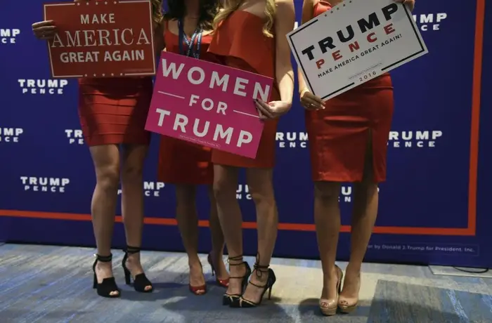 Tre kvinner i røde kjoler og hæler holder pro-Trump-skilt, inkludert «Make America Great Again»; og "Kvinner for Trump" ved et politisk arrangement