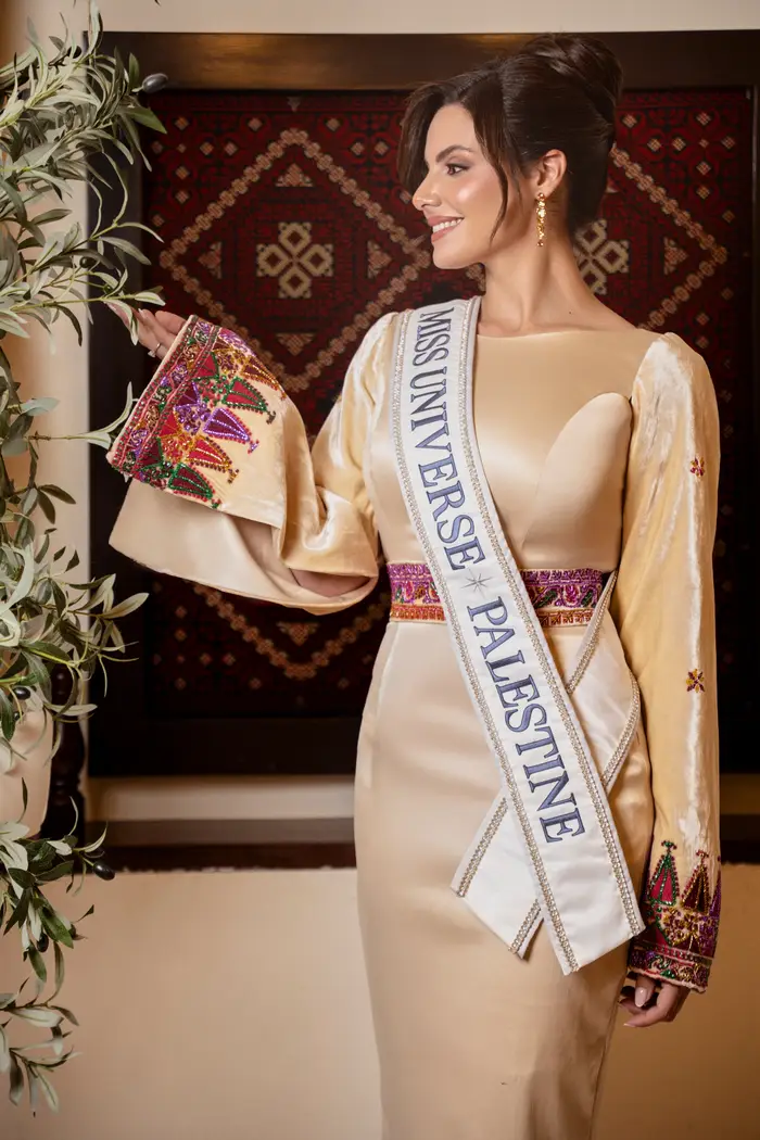 Kvinne iført en Miss Universe Palestine-sash, i en gulvlang kjole med intrikate ermer, stående innendørs nær en plante