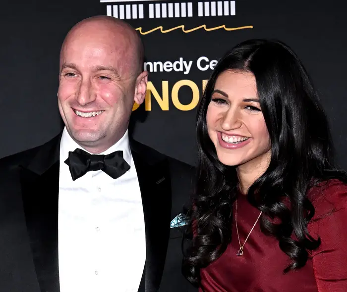 To personer poserer på et formelt arrangementsteppe. Kvinnen har på seg en langermet, figursydd kjole, og mannen har på seg en klassisk smoking med sløyfe