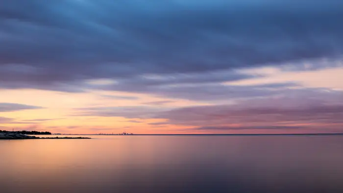 Stille hav under en vid himmel med lange, pisket skyer ved solnedgang. Fjern kystlinje synlig i horisonten