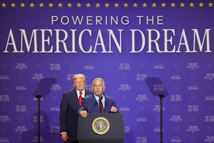 To menn i dress står på et podium med blått bakteppe og leser «Powering the American Dream»; ved et politisk arrangement