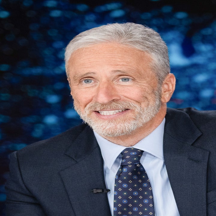 Jon Stewart i dress sitter ved en nyhetsskranke og smiler