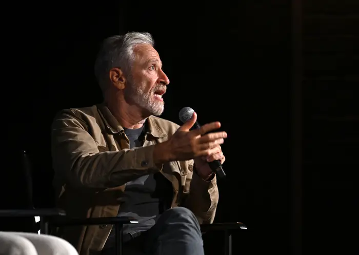 Jon Stewart snakker i en mikrofon mens han gestikulerer med hånden, sittende på scenen i en uformell jakke