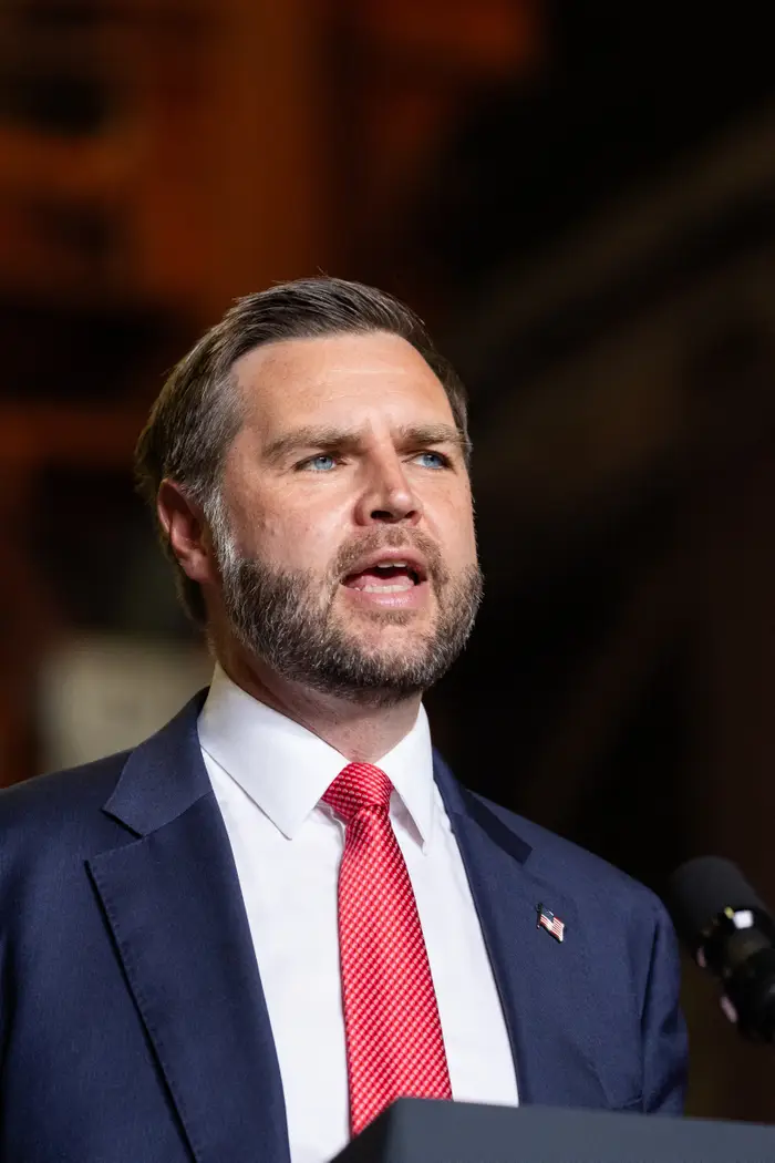 En mann i dress og rødt slips snakker ved et podium med en mikrofon, formidler en tale eller kommer med en kunngjøring