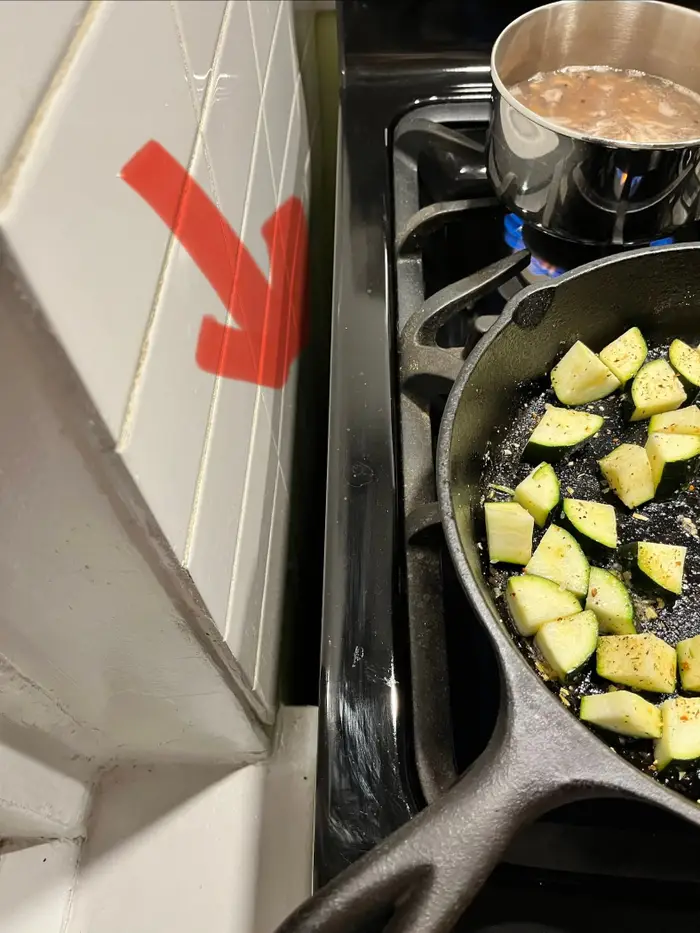 Zucchinibiter som sauteres i en panne på en komfyr med en gryte ved siden av, nær en vegg merket med en stor rød pil