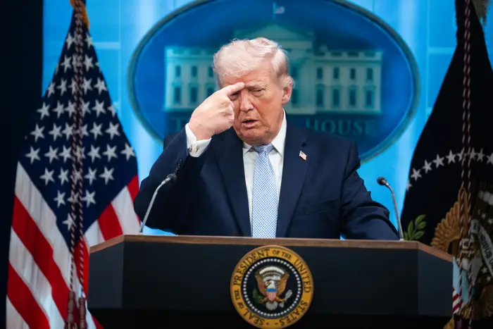 En person i dress gestikulerer nær pannen ved et podium med amerikanske flagg og et bakteppe som ligner Det hvite hus