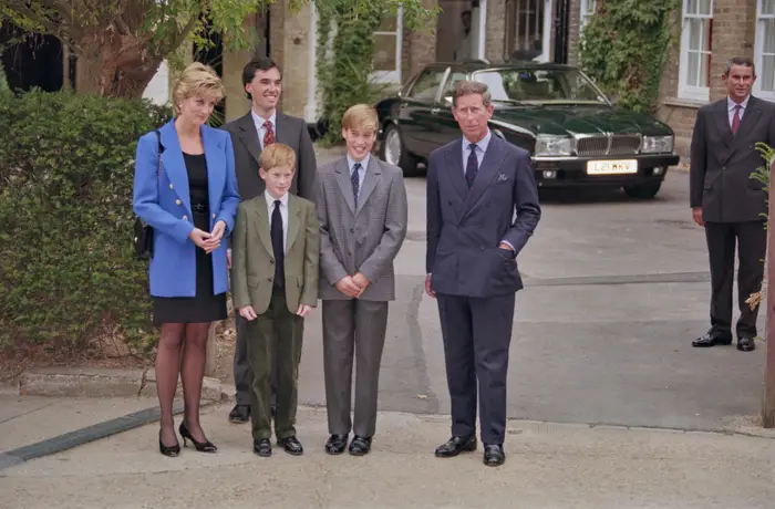 En gruppe velkledde individer, inkludert en kvinne i blå blazer og menn i dress, står utendørs nær en parkert klassisk bil