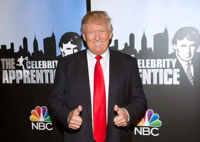 Person i dress med rødt slips gir tommelen opp på "The Celebrity Apprentice" begivenhet, med NBC-logoer i sikte
