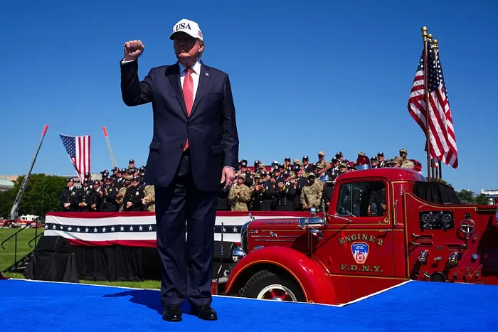 Mann i dress og USA-caps hever neven på scenen med brannbil, mengde uniformerte mennesker og flagg i bakgrunnen. Patriotisk begivenhet