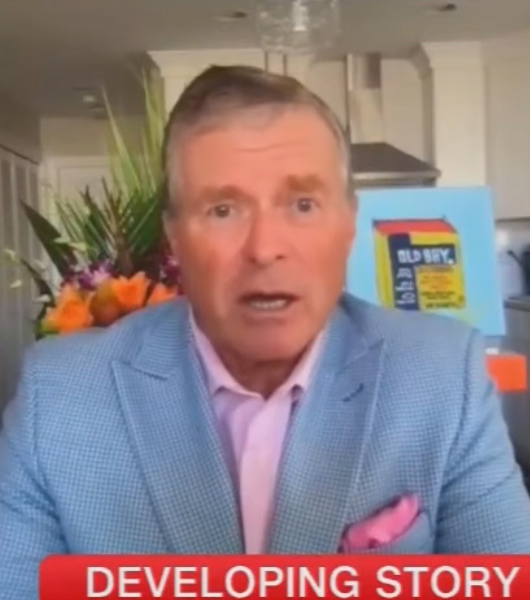 En person i blå dress og rosa skjorte snakker i et rom med blomster og en bok med tittelen "GAMMEL SPION" synlig i bakgrunnen. Nyhetstickeren lyder 