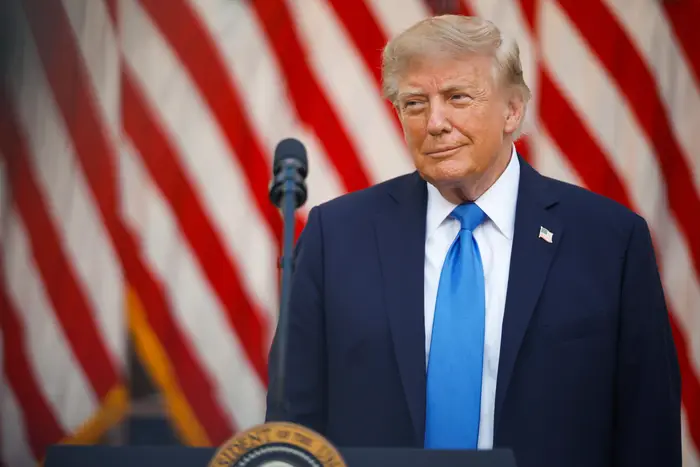 En mann i dress og blått slips står ved et podium med et amerikansk flagg i bakgrunnen