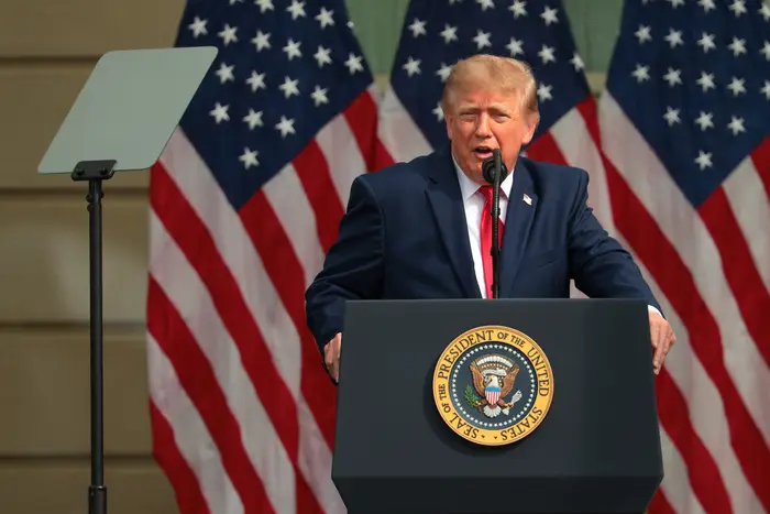 Donald Trump i dress taler ved et podium med presidentsegl, omgitt av flagg. Foredragsarrangement