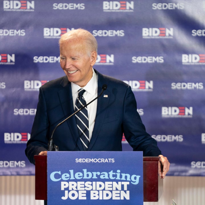 Mann i dress taler ved et podium med skilt: «Celebrating President Joe Biden». Bakgrunnsbannere viser «SODEMS» og «BIDEN»-logoer