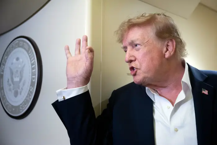 Person som gestikulerer ved siden av det amerikanske presidentseglet på en vegg, iført dress og hvit skjorte med åpen krage, og ser ut til å snakke