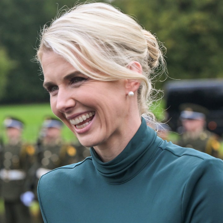 Person som smiler utendørs, kledd i et antrekk med høy hals. En gruppe uniformerte individer er synlig i den uskarpe bakgrunnen