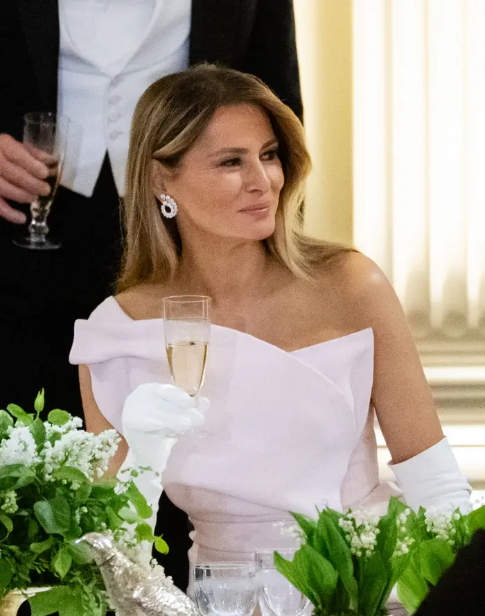 En kvinne på et elegant arrangement holder et champagneglass, iført en off-shoulder kjole, hvite hansker og glitrende øredobber, omgitt av grøntområder