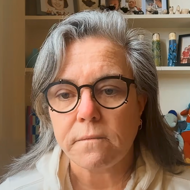 Person med briller og middels langt hår, iført en lys topp, sitter innendørs med hyller som viser innrammede bilder og leker bak dem