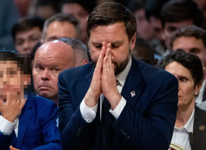 En mann og en ung gutt i formell antrekk sitter blant en folkemengde; mannen har hender i bønn, og gutten legger haken på hånden og ser tankefull ut