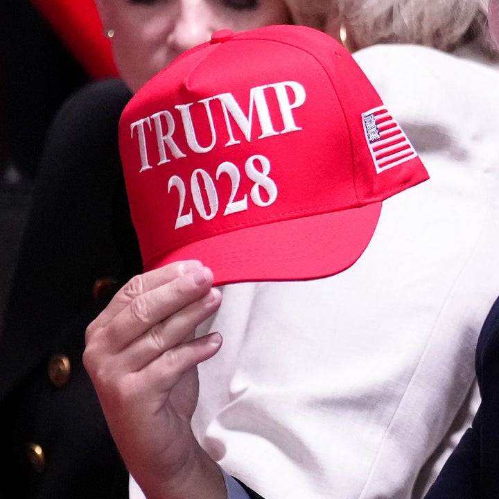 Mann i dress sitter, smiler og holder en rød "Trump 2028"-hette under et arrangement