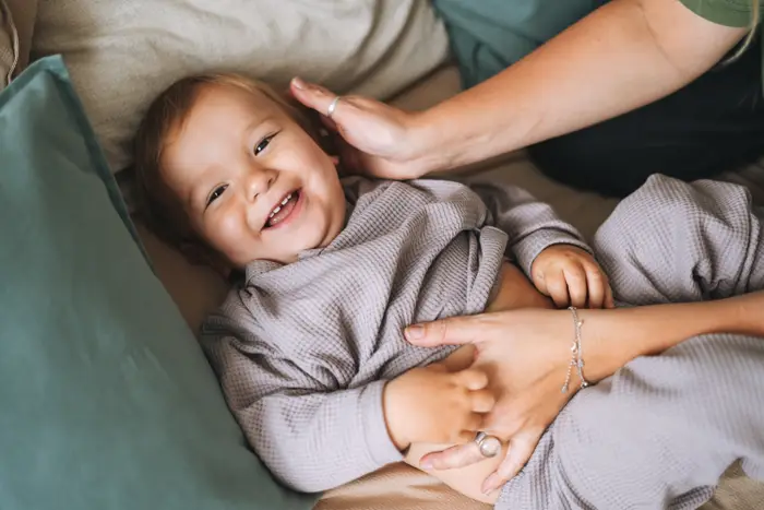Baby smiler mens han ligger på en seng, lekent samhandler med en voksens hender