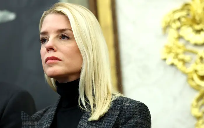 Person med langt, rett hår og et seriøst uttrykk, iført en høyhalset topp og blazer, står i en formell setting