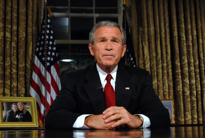 George W. Bush i dress satt ved skrivebordet og snakket med et alvorlig uttrykk. Amerikanske flagg i bakgrunnen, innrammede bilder på skrivebordet