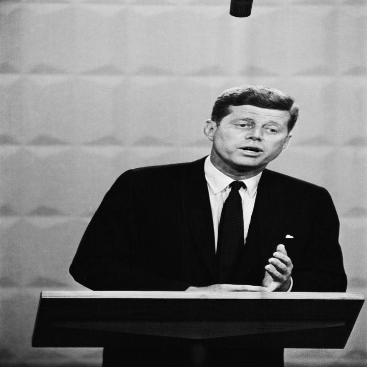 JFK i dress taler ved et podium og henvender seg til et publikum eller kameraer i en formell setting